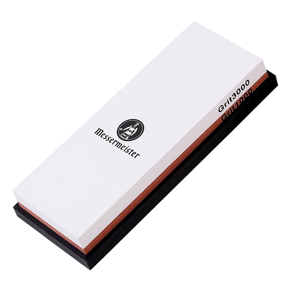 Messermeister 1000 & 3000 grit maintenance Stone is a dual sided rectangle 8 inch x 3 inch x 1 inch. The top layer is white with black "Messermeister" logo. "Grit3000" labeled in black along the top layer.  Bottom layer is reddish brown with "Grit1000" labeled. The item sits in a rectangular black silicone base. Image tilted up and to the right on a white background.