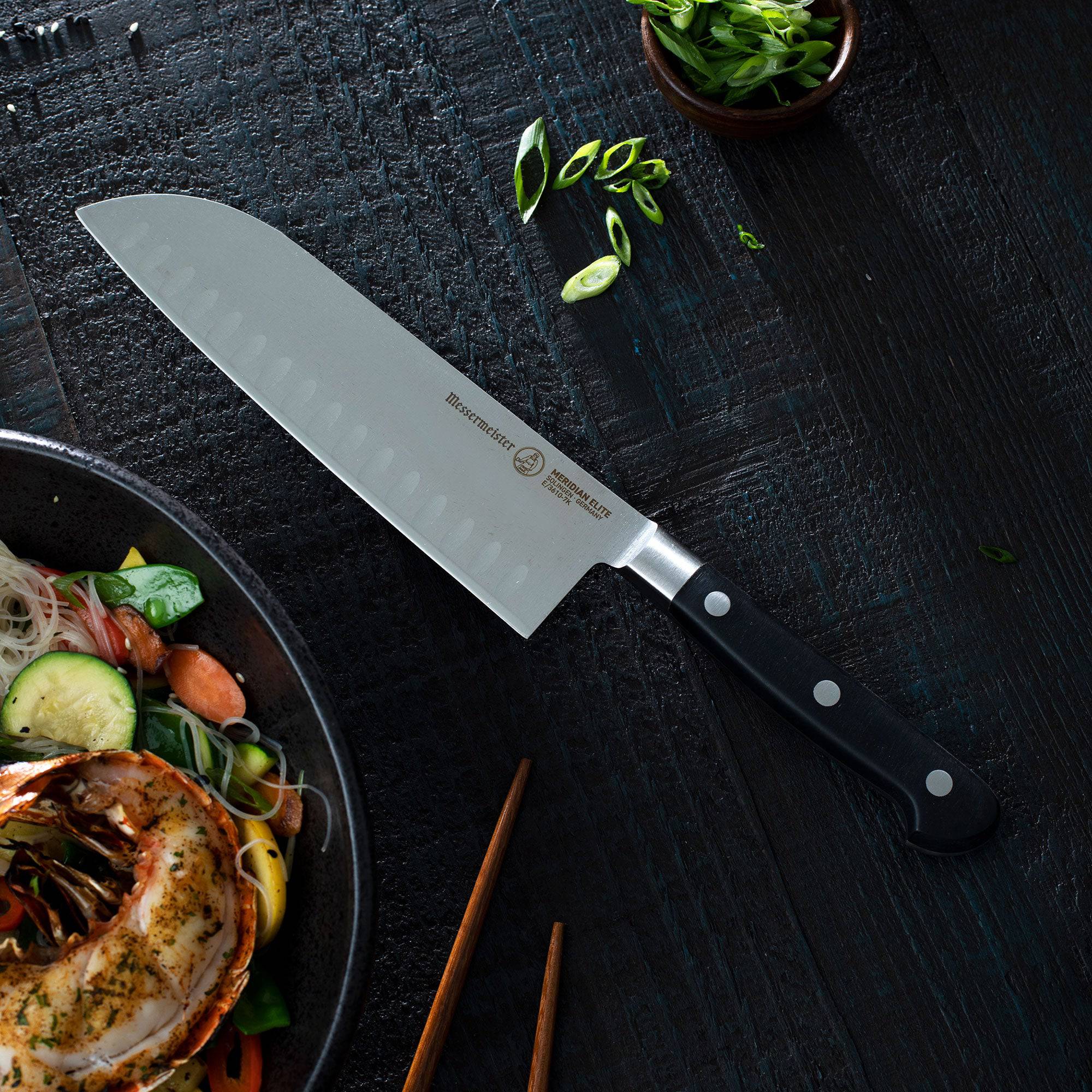 A 7 inch Meridian Elite Kullens Santoku Knife laying on its side next to a bowl containing grilled lobster tail and rice noodles with vegetables. 