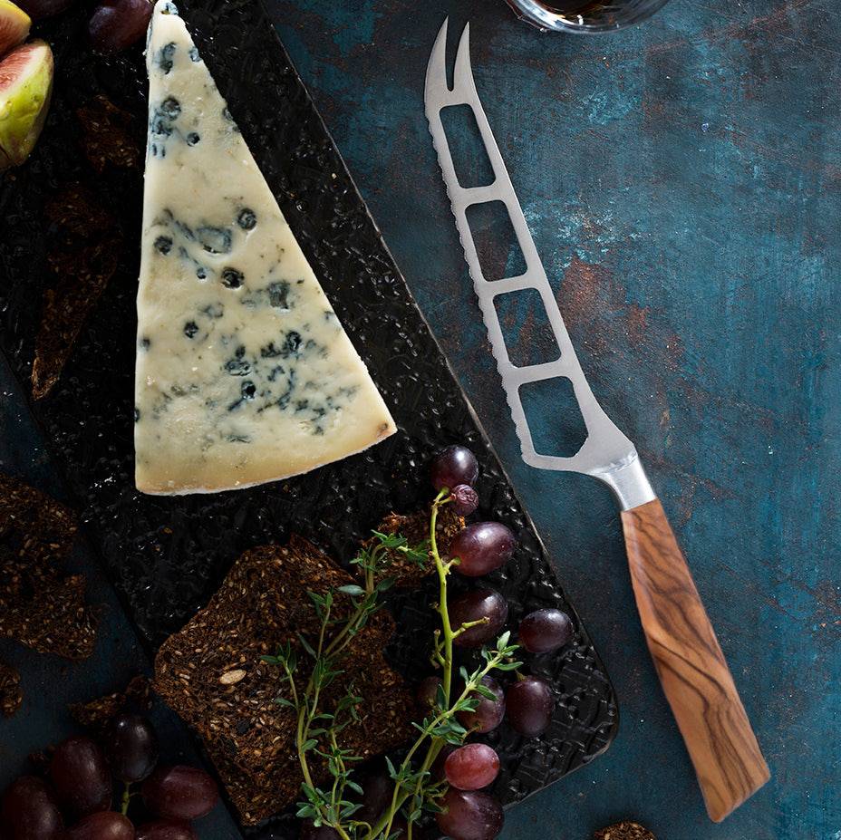 The Oliva Elite Cheese and Tomato Knife with a stainless steel blade with holes and two prongs at the tip with olive wood handle lying next to a wedge of blue cheese and grapes