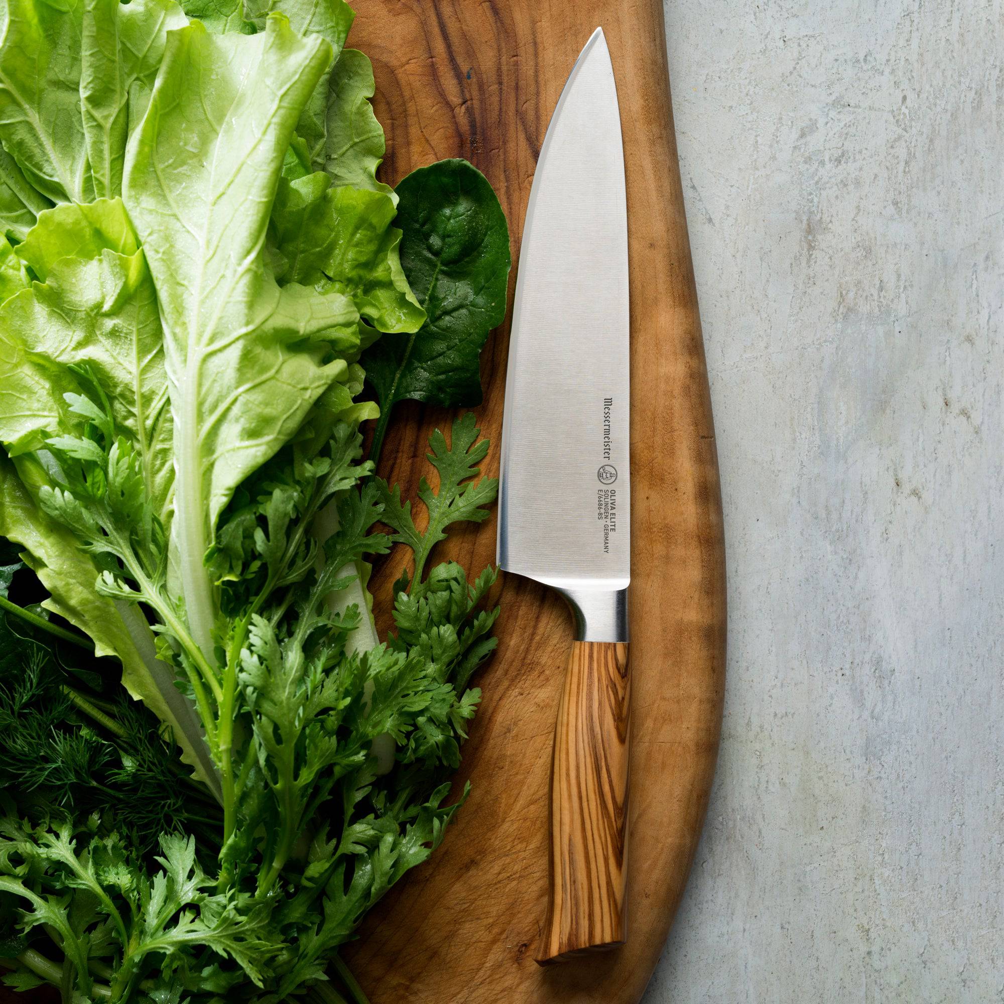 Photo of the Oliva Elite Chef's Knife resting on cutting board next to a pile of leafy greens