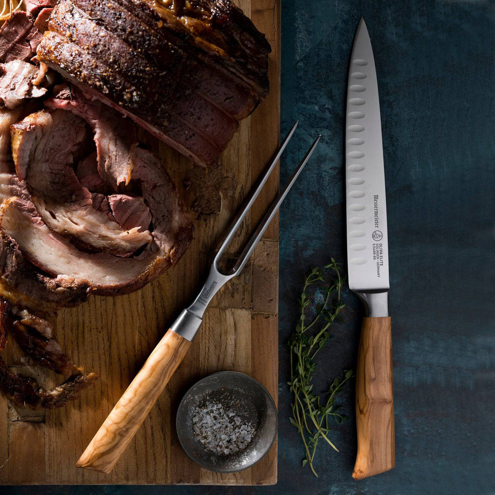The Oliva Elite Kullens Carving Set shown resting next to a roast beef on a wooden cutting board with a small dish of salt and herbs