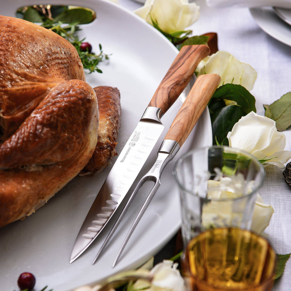 A close up of the Oliva Elite Kullens Carving Set shown resting next to a roast turkey on a large platter with herbs and flowers 