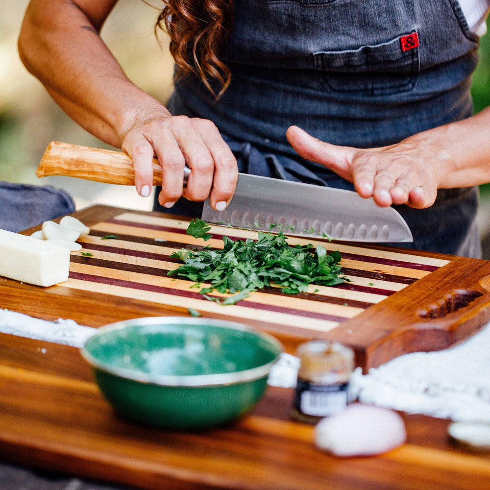 A person is chopping herbs on a wooden cutting board the Oliva Elite Santoku knife.