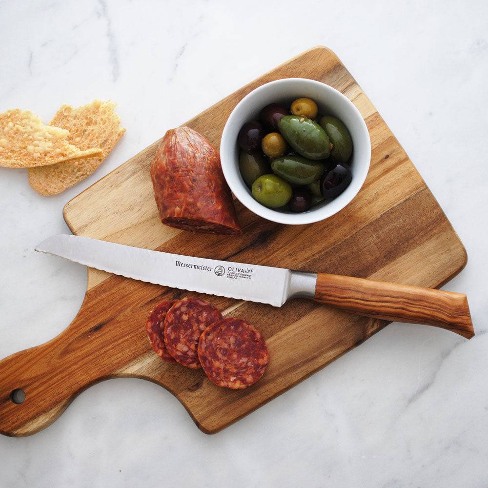 The Oliva Elite 6 inch Reverse Scallop Utility Knife with olive wood handle and a silver stainless steel blade shown resting on wooden cutting board with salami and olives