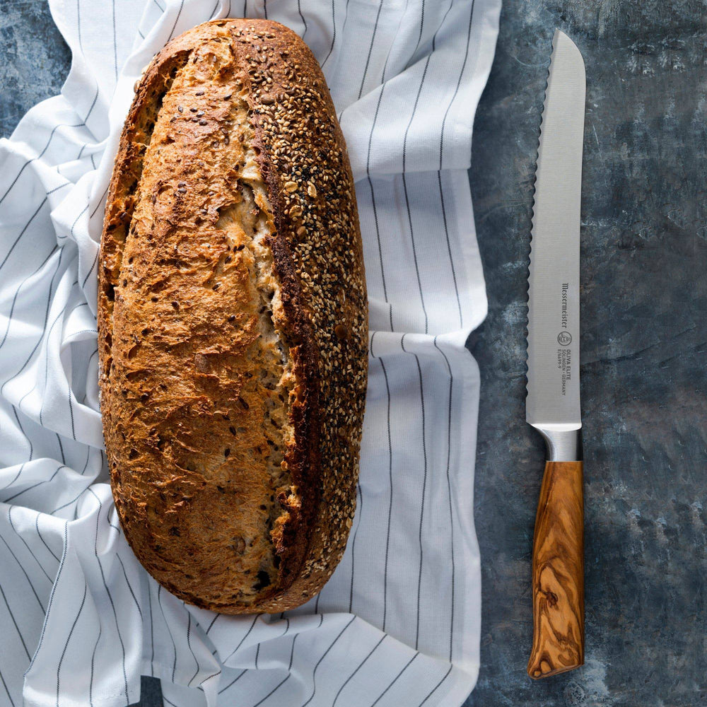 The Oliva Elite 9 inch serrated bread knife with silver stainless steel blade and olive wood handle shown lying next to a seeded loaf of bread that is on top of a tea towel