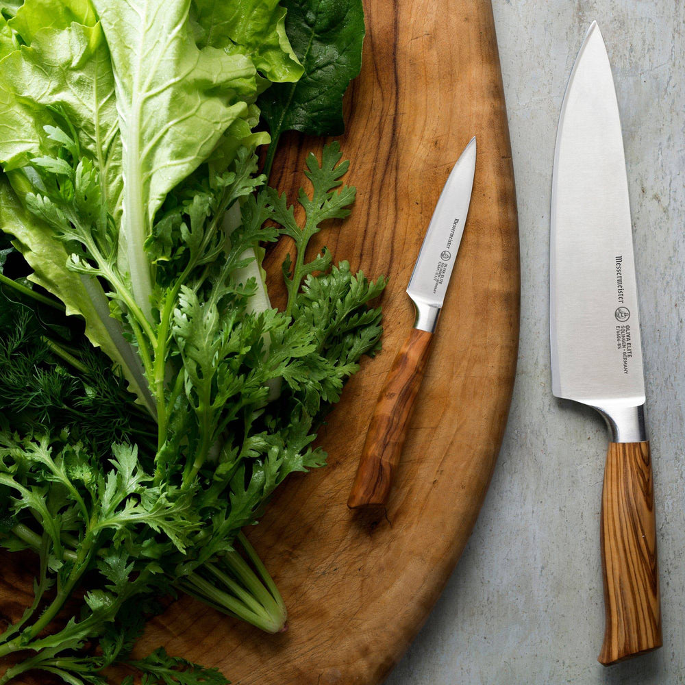 The Oliva Elite Chef and Parer set with the 8 inch Chef's Knife and the 3.5 inch Paring Knife both with steel blades and olive wood handles shown resting next to leafy greens on a wood cutting board